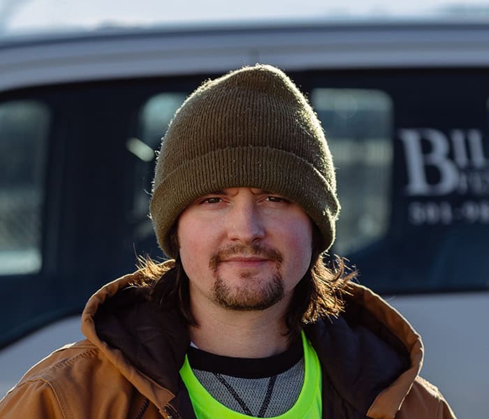 Brian Sharp with long brown hair and a goatee wears a brown beanie, a neon green shirt, and a brown jacket, standing outdoors in front of a white vehicle.
