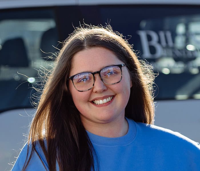 Bryanna Cook with long brown hair and glasses smiles while standing outside in front of a white vehicle. She is wearing a blue sweatshirt and sunlight is shining on her face.