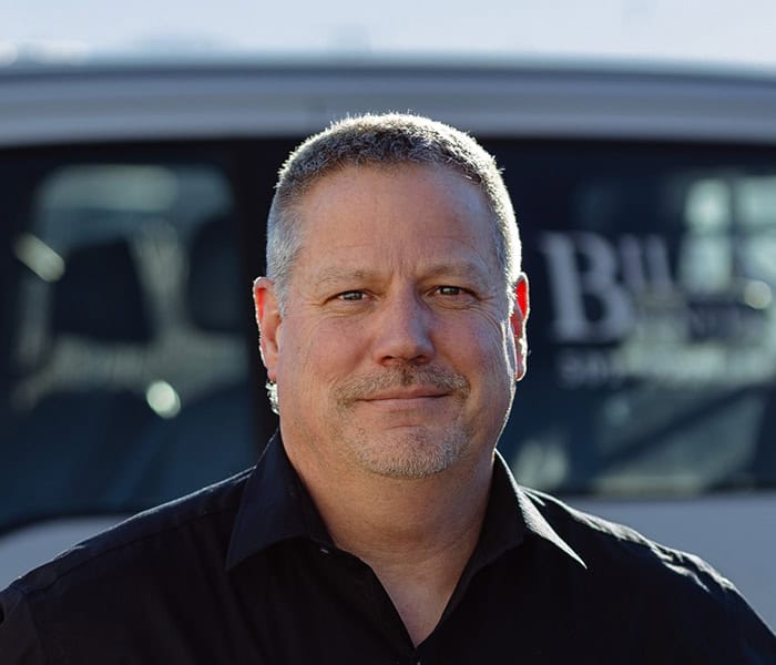 Chris Brown with short gray hair, wearing a black collared shirt, stands outside in front of a vehicle with blurred writing on the window. He is looking at the camera and smiling slightly.