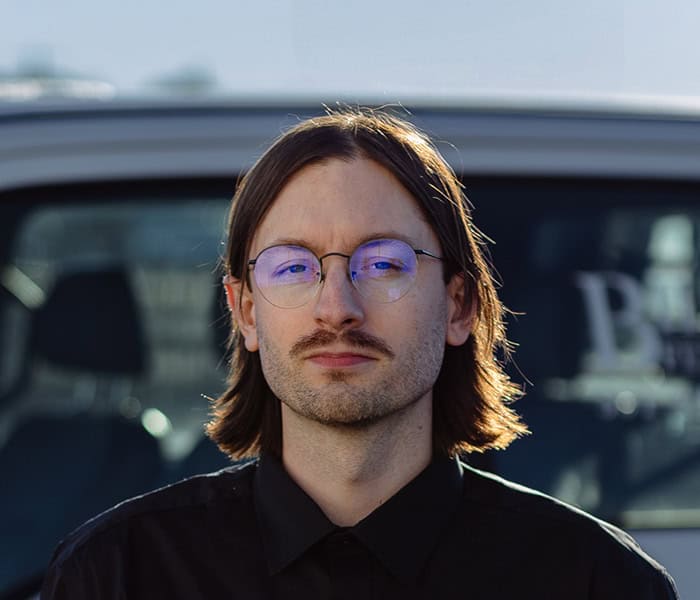 Dalton Pabst with shoulder-length brown hair, glasses, and a mustache stands in front of a vehicle, wearing a black shirt and looking directly at the camera with a neutral expression.