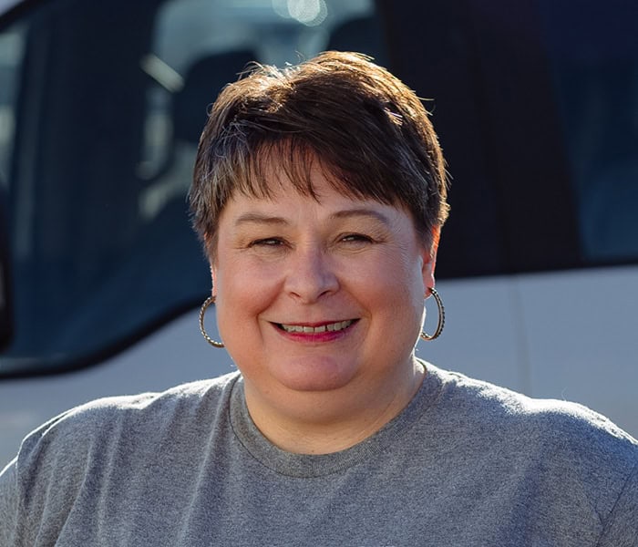 Erin Glass with short brown hair, wearing hoop earrings and a gray t-shirt, smiles at the camera while standing outside in front of a white vehicle.