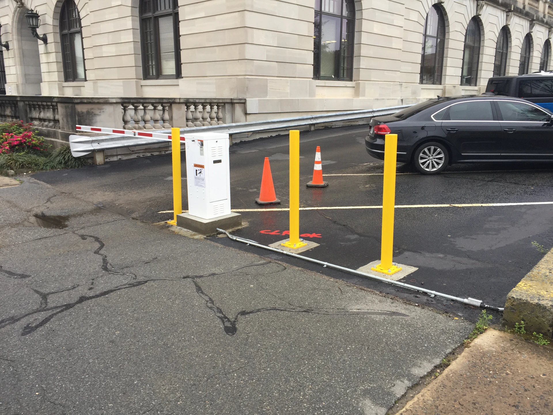 A parking lot entrance with three yellow bollards, two orange traffic cones, a white control box, and a red-and-white barrier arm, next to a parked black car by a stone building.