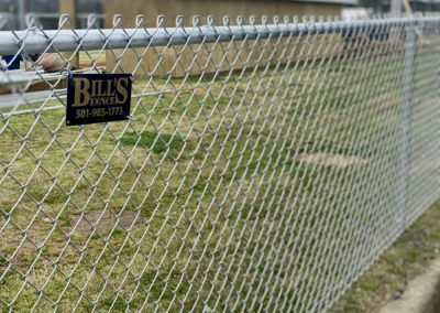 A close-up view of a chain-link fence with a sign reading BILLS FENCE 501-985-1773, enclosing a grassy area near buildings in the background.