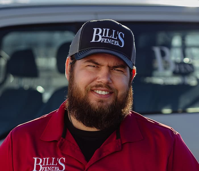 Jesse Adams wearing a “BILLS FENCE” cap and a red polo shirt stands in front of a vehicle with the same company logo visible in the background. He is smiling and facing the camera.