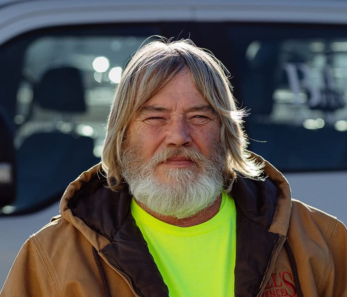 Jimmy Champlin with long gray hair and a full beard stands outdoors, wearing a brown jacket over a bright yellow shirt, with a vehicle visible in the blurred background.