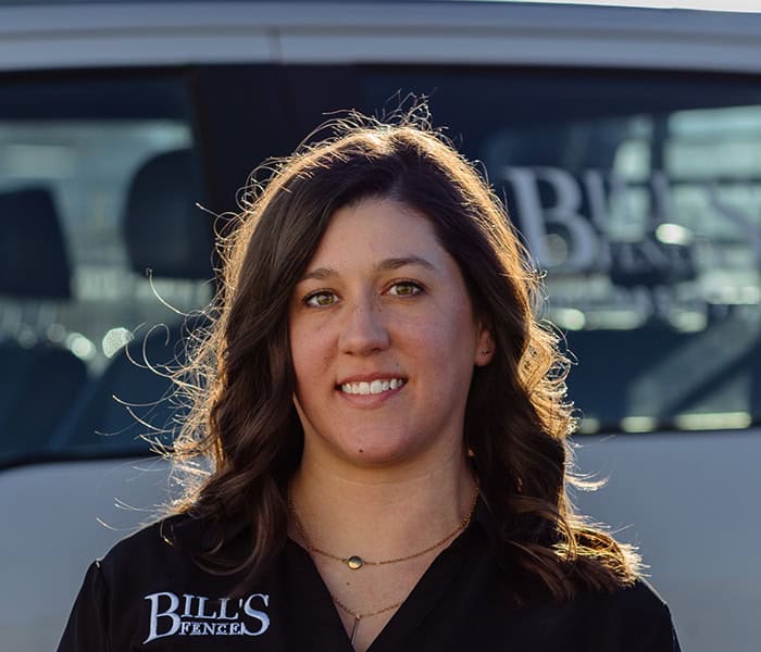 Mary Bridges with wavy brown hair wearing a black shirt that says BILLS FENCE stands outdoors, smiling, with a white vehicle in the background.