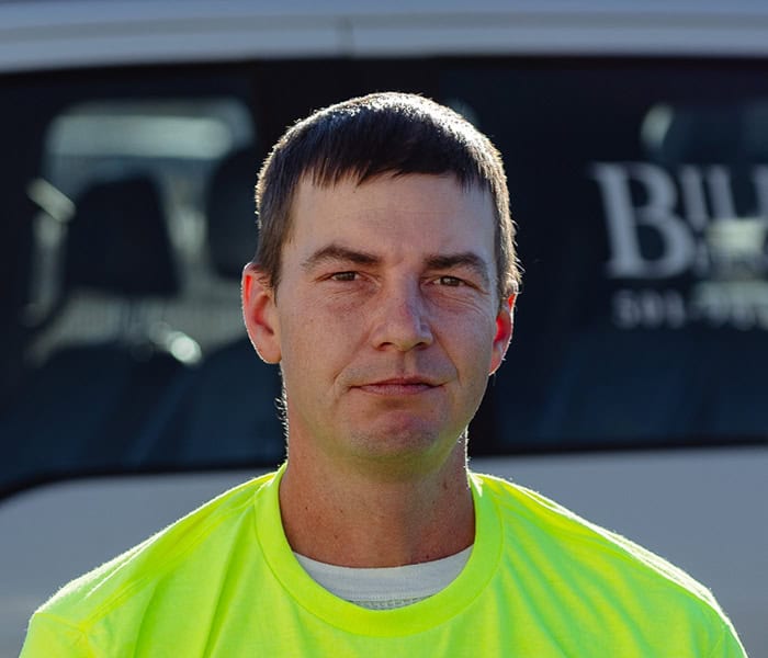 Matt Mims with short brown hair and a neutral expression wears a bright yellow shirt, standing in front of a white vehicle with partially visible text on its window.