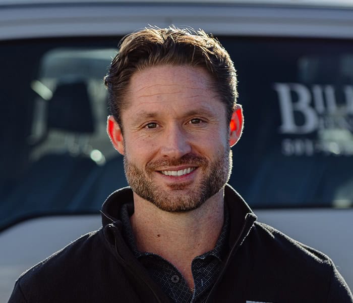Michael Pabst with short, wavy brown hair and a trimmed beard stands outdoors in front of a white vehicle. He is wearing a dark jacket over a collared shirt. Sunlight highlights the edge of his face and ears.