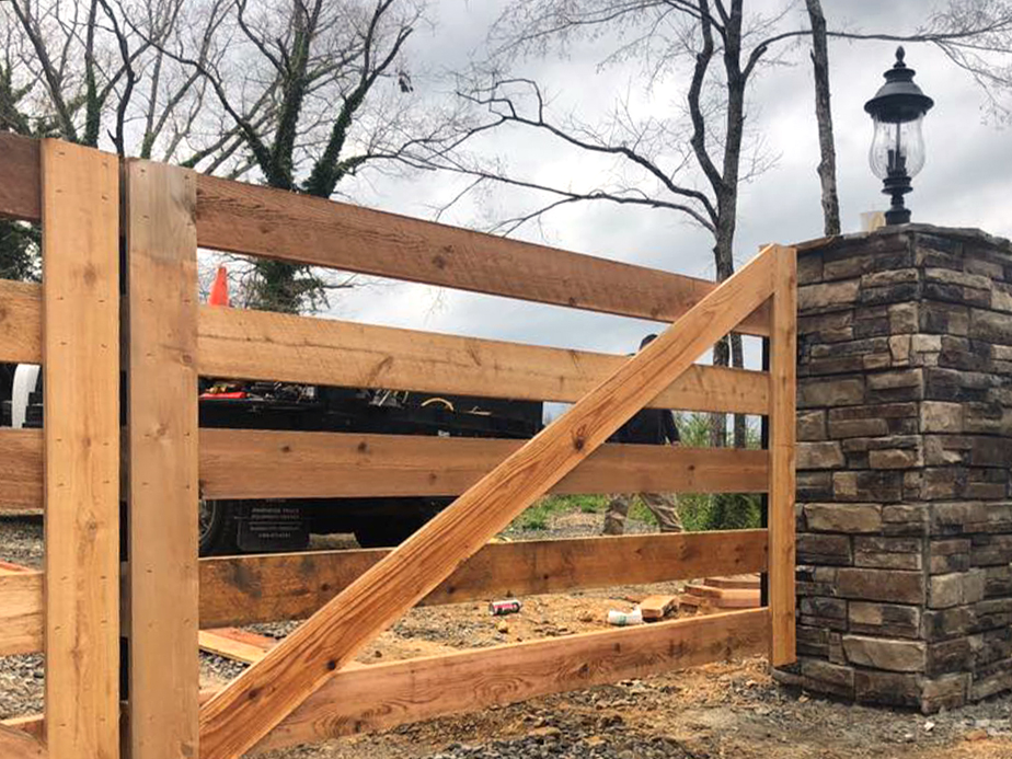 A wooden gate with diagonal support is attached to a stone pillar topped with a black lantern, set outdoors with trees and cloudy sky in the background.