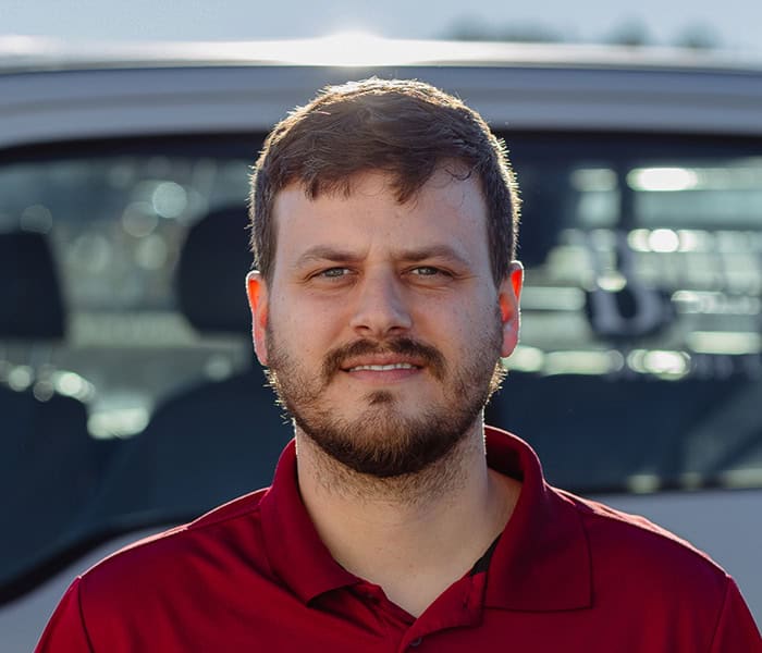 A man with short brown hair and a beard, wearing a red collared shirt, stands outside in front of a white vehicle, looking directly at the camera.