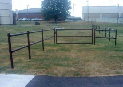 A short iron fence stands on grass, but it abruptly ends without enclosing anything, making it nonfunctional; buildings and parked cars are visible in the background.