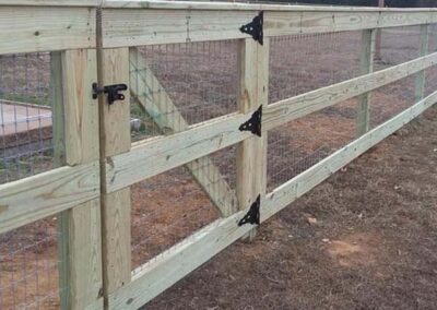 A wooden and wire mesh fence with a gate, featuring black metal hinges and latch, stands on dry grass with a field in the background.