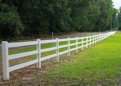 A long white wooden fence runs alongside green grass and a dirt path, with dense trees and foliage in the background under a clear sky.