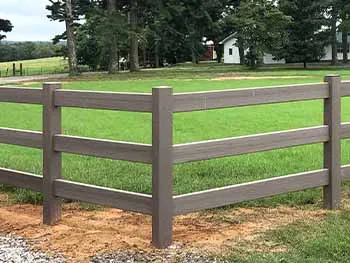 A three-rail brown vinyl fence surrounds a grassy area near a gravel path, with trees and a white house visible in the background.