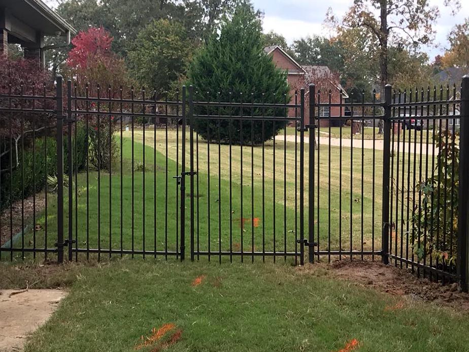 A black metal fence with a gate encloses a grassy yard. Orange markings are visible near the ground, and a paved path leads up to the fence. Trees and a house are in the background.