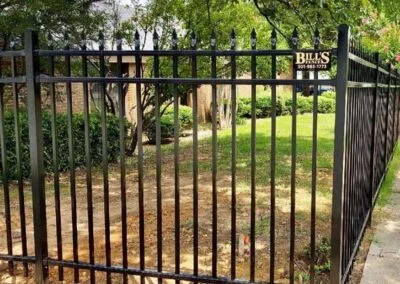 A tall black metal fence with vertical bars encloses a yard with green grass and trees. The fence has a sign reading BILLS with a phone number. The ground outside the fence is bare soil.