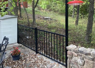 A black metal fence separates a stone patio with a potted red flower from a grassy, wooded area. A red bird feeder hangs above the fence on a black pole. Some fallen leaves are scattered on the patio.