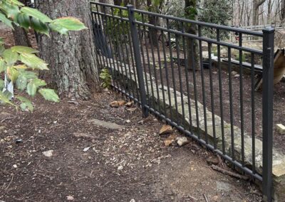 A black metal fence is built over a stone border between trees in a wooded area. The uneven ground slopes downward, and the fence follows the incline, creating a gap beneath it.