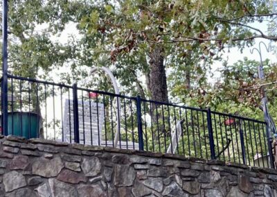 A metal fence sits atop a stone wall, with leafy tree branches overhead. Behind the fence are outdoor furniture, including stacked chairs and a green barrel. White cables or hoses hang over the fence.