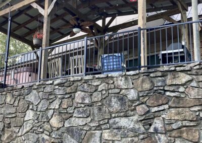 A stone wall supports a raised patio with a wooden pergola, black metal railing, outdoor chairs, a hanging plant, ceiling fans, and a barbecue grill.