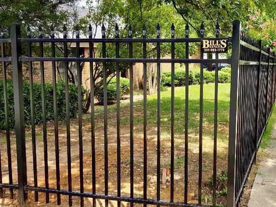 A black metal fence with decorative points surrounds a green lawn and shrubs. A sign on the fence reads BILLS with a phone number. Trees and part of a brick building are visible in the background.