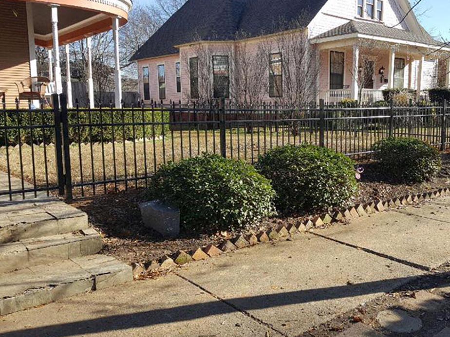 A black metal fence surrounds a yard with trimmed bushes and patchy grass in front of two large, old-style houses with wide porches and gable roofs. Stone steps lead up to the fence from the sidewalk.