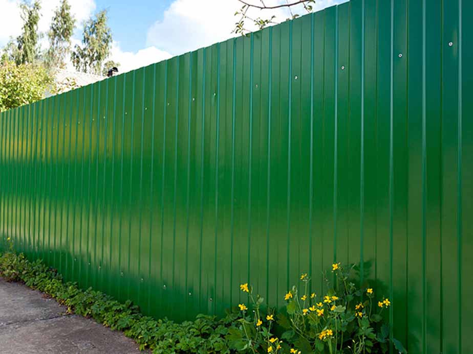 A tall, green metal fence runs alongside a paved path, with yellow wildflowers and green plants growing at its base, against a background of trees and blue sky with clouds.