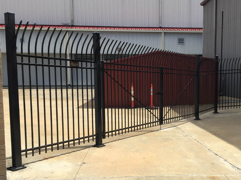 A black metal security fence with curved spikes on top encloses a concrete area. The fence features a double gate, and behind it is a large red metal shed and two orange traffic bollards.