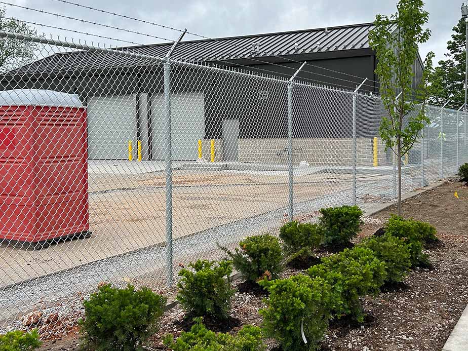 A construction site enclosed by a chain-link fence topped with barbed wire, with a red portable toilet outside and small green shrubs planted along the sidewalk.