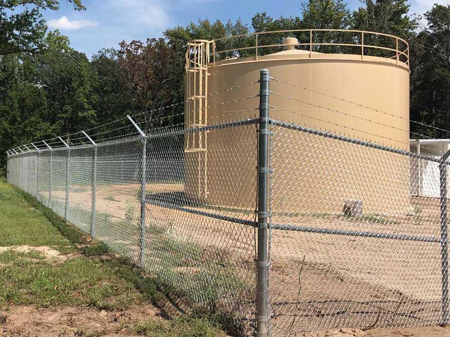 A large tan cylindrical water storage tank is enclosed by a chain-link fence topped with barbed wire, surrounded by trees and grass under a partly cloudy sky.