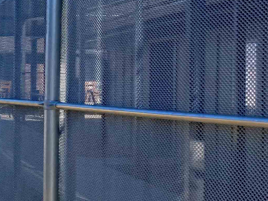 A close-up view of a metal mesh fence with horizontal and vertical support bars, partially obscuring the blurry background behind it.