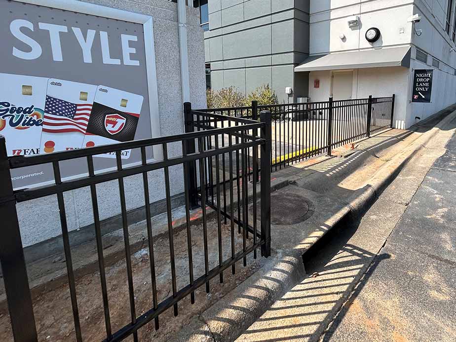 A fenced area blocks the sidewalk next to a drive-thru lane, forcing pedestrians to walk on the road. A signboard with images of gift cards is visible on the building wall to the left.