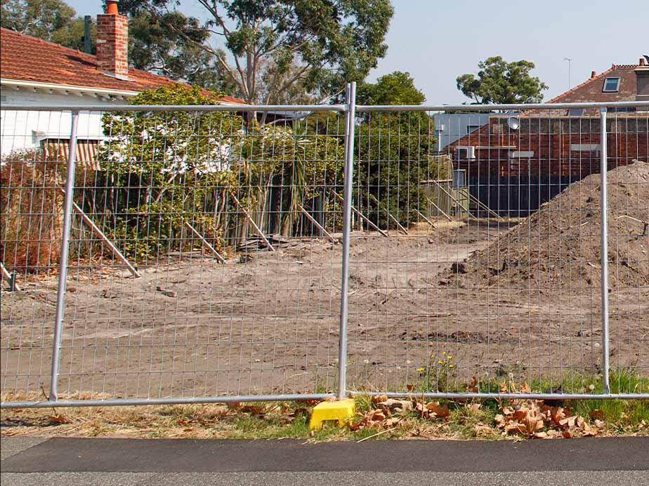 A metal fence blocks access to a cleared, dirt-filled lot between houses, with a pile of soil and scattered leaves in front. Trees and parts of neighboring homes are visible in the background.