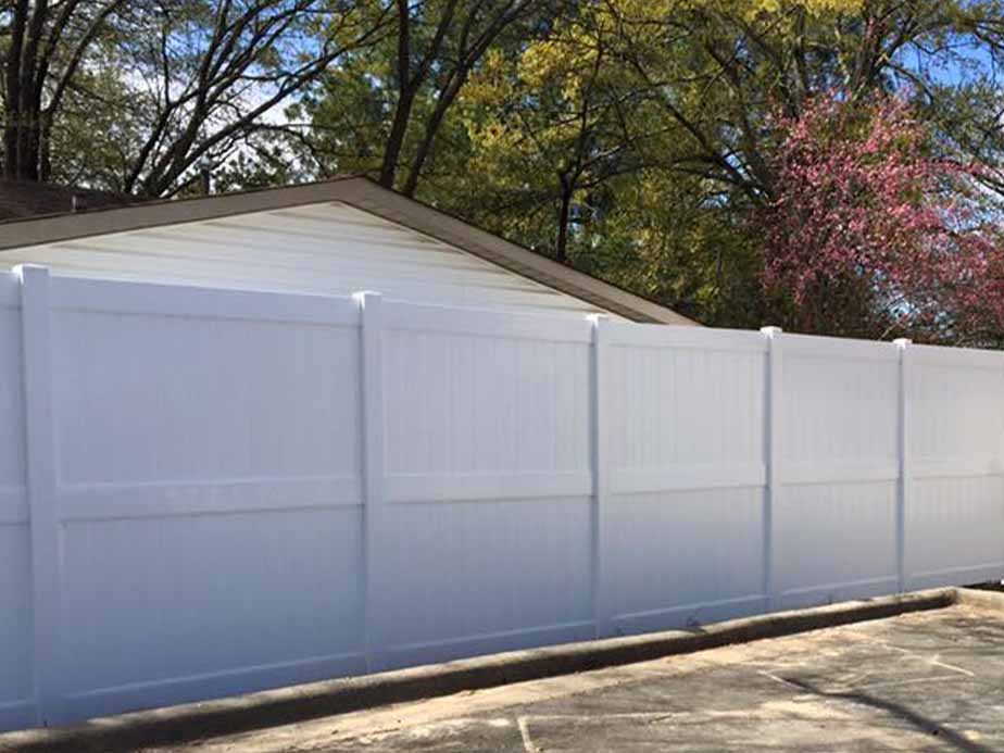 A tall white vinyl privacy fence stands in front of a house with a sloped roof, surrounded by trees with green and pink foliage. The scene is bright and outdoor pavement is visible in the foreground.