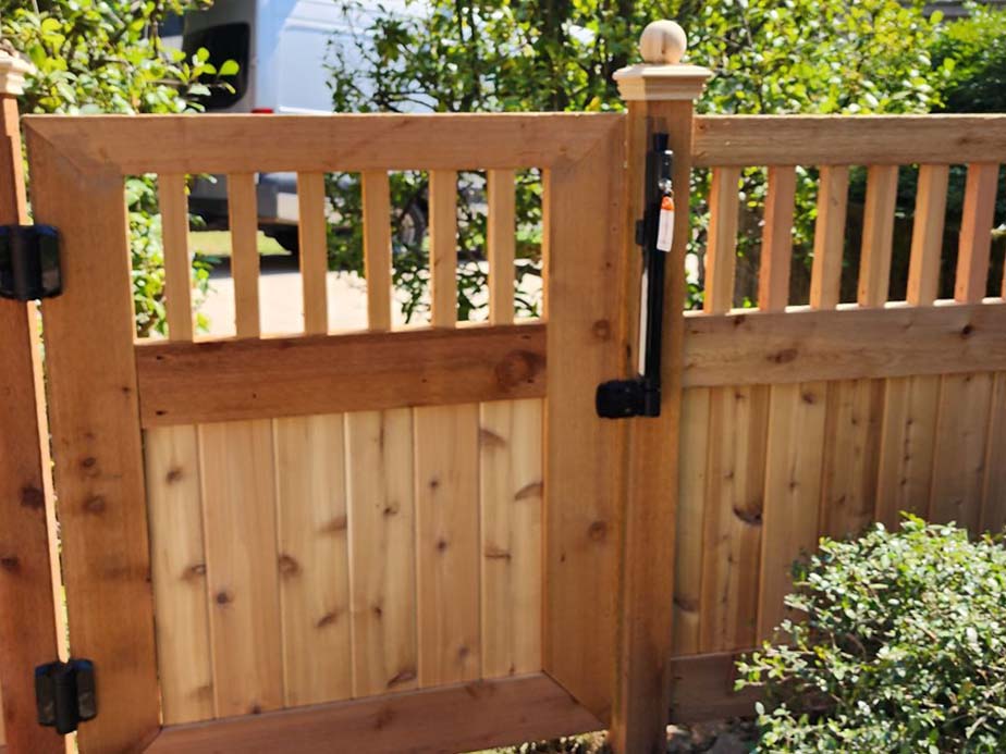 A close-up of a wooden garden gate with vertical slats and black metal hinges, surrounded by green bushes and foliage, with part of a white vehicle visible in the background.