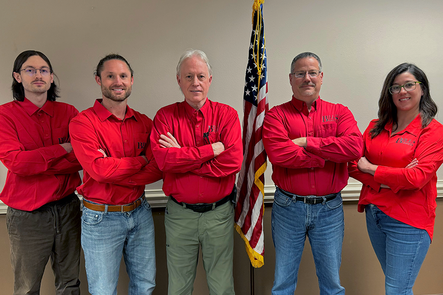 Five people in matching red shirts stand in a row with arms crossed, posing in front of an American flag against a beige wall.