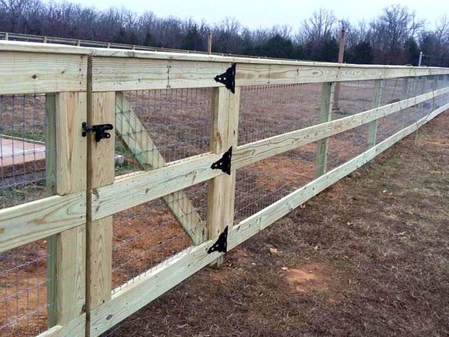 A long wooden fence with wire mesh stretches across a grassy area. The fence has metal hinges and a small black latch on a gate section. Leafless trees are visible in the background under a cloudy sky.