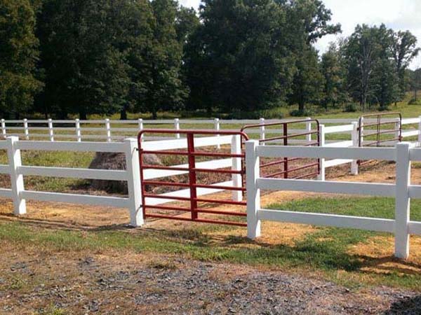 A white fence with a red gate is positioned over a shallow ditch, but the gate does not block access since the fence ends at the edge of the ditch, making it easy to walk around. Trees and grass are visible in the background.