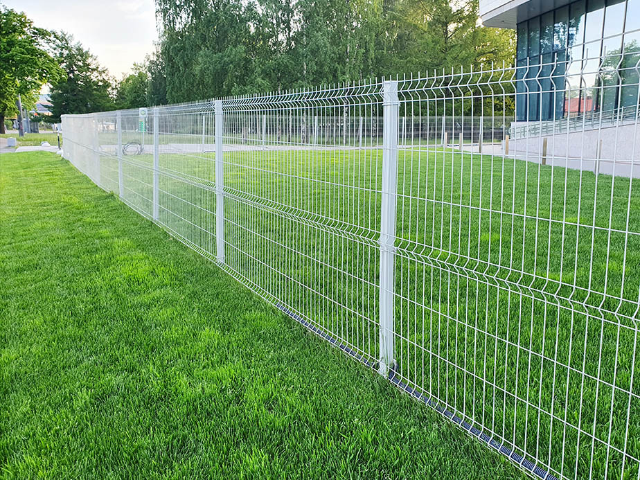 A metal wire fence runs across a well-maintained grassy lawn, with trees and a modern glass building in the background. The scene appears to be in an urban or park-like setting.