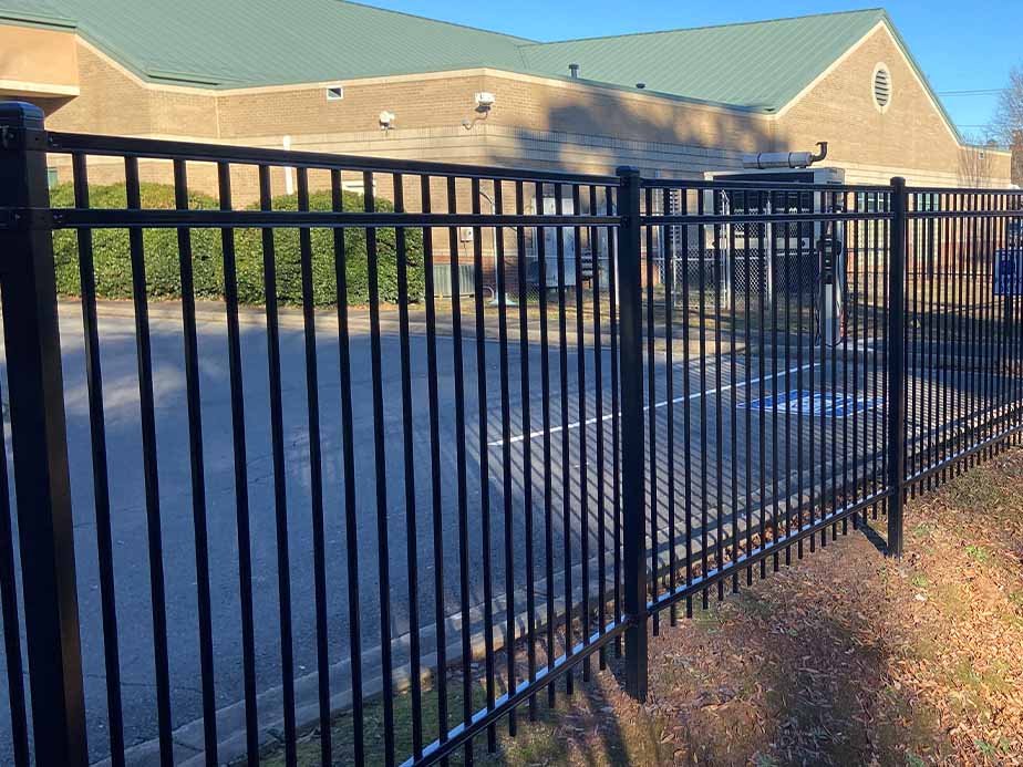 Black metal fence in front of a brick building with a green roof. The fence encloses a parking lot with a visible accessible parking space. Bushes line the side of the building.