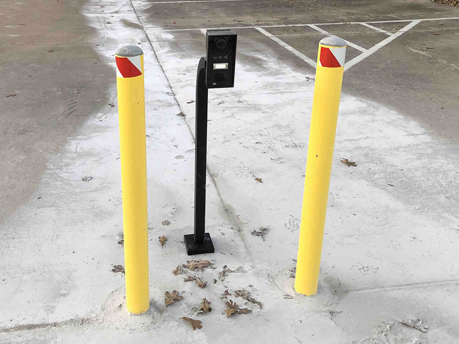 A black parking lot ticket machine stands between two yellow bollards with red reflective tape, on a concrete surface with scattered brown leaves. White parking lines are visible in the background.