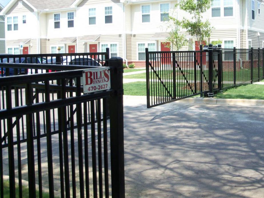 Black iron security gates surround the entrance to a residential apartment complex with beige buildings and red doors. A sign on the gate reads Bills Fence with a phone number. The gates are open on a sunny day.
