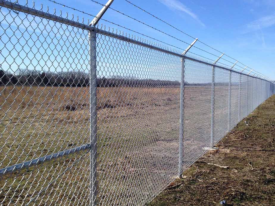 A tall chain-link fence topped with barbed wire runs along a grassy field under a blue sky, serving as a boundary or security barrier.