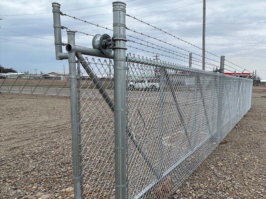 A tall chain-link security fence topped with barbed wire runs along a gravel lot under a cloudy sky. The fence features a sturdy sliding gate supported by metal posts and rollers.