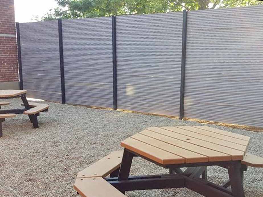 A gravel-covered outdoor area with wooden picnic tables and a tall corrugated metal privacy fence with black posts, next to a red brick wall. Trees are visible above the fence.