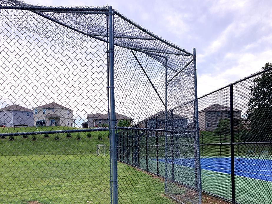 A chain-link fence separates a grassy field from blue tennis courts. Houses are visible in the background on a grassy hill under a partly cloudy sky.