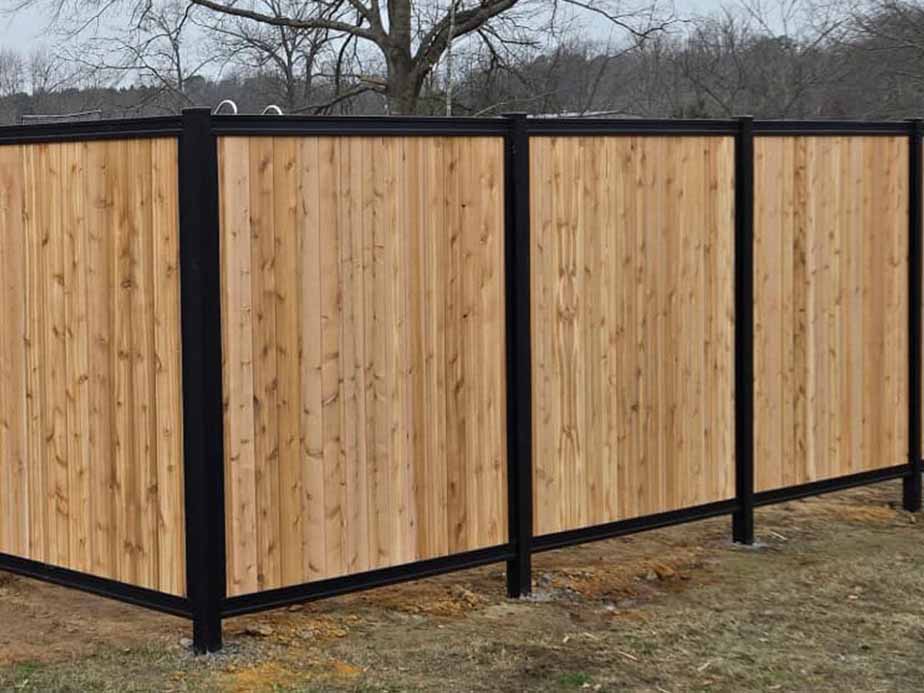 A wooden privacy fence with vertical planks framed by black metal posts stands on a grassy area with bare trees and cloudy skies in the background.