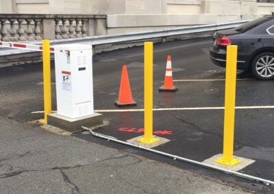 Four yellow bollards surround a white control box near a parking lot entrance. Two orange traffic cones and a car are in the background, with CLEAR painted in red on the asphalt.