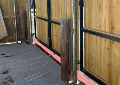 A wooden fence with metal posts, a concrete ground, and a single brown bollard in front of the gate. Sunlight casts shadows on the ground.