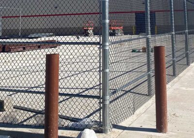 A chain-link fence encloses a concrete area with construction materials and two orange boom lifts in the background. Two brown metal posts stand near the fence on the sidewalk.